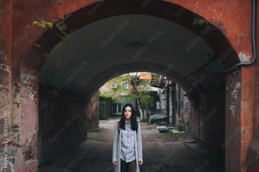A young sad girl in a gray cardigan and a shirt stands in the arch of an old slum yard and looks at the viewer. The concept of poverty, decline and lack of prospects.