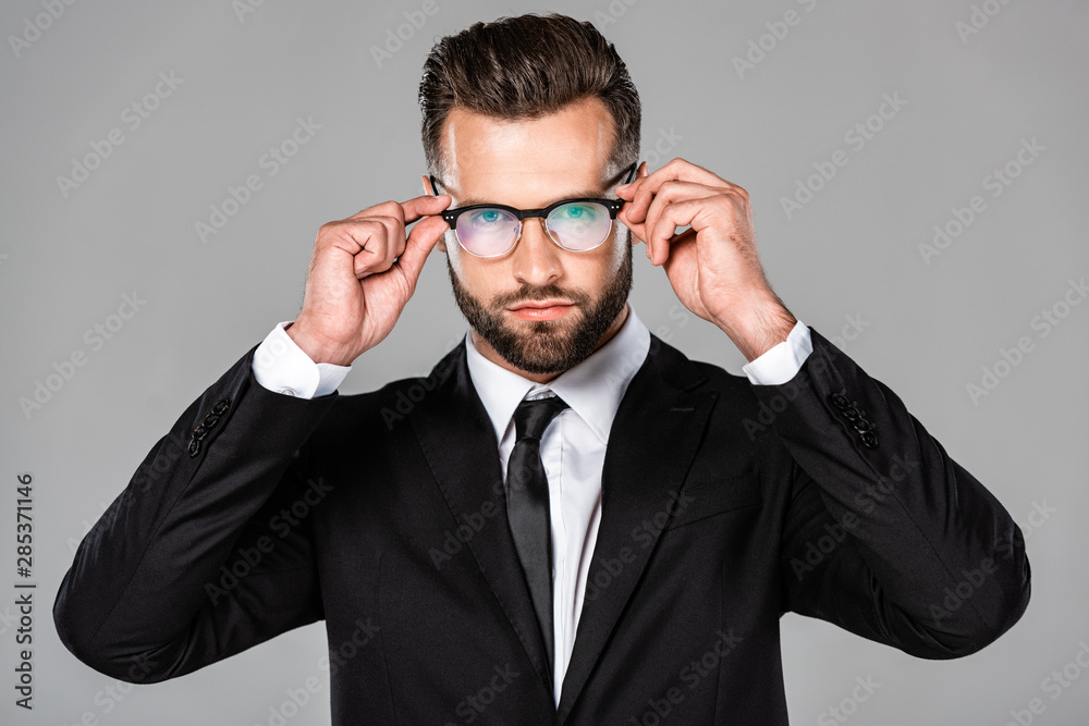 successful businessman in black suit touching glasses isolated on grey successful businessman in black suit touching glasses isolated on grey
