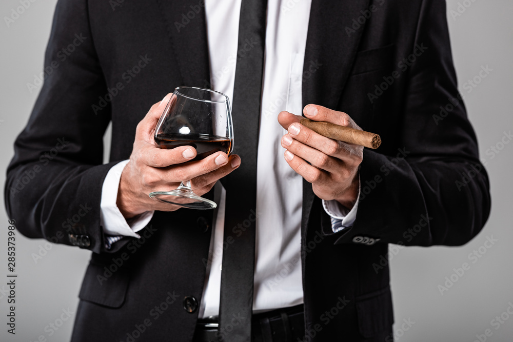 partial view of businessman in black suit holding glass with whiskey and cigar isolated on grey partial view of businessman in black suit holding glass with whiskey and cigar isolated on grey