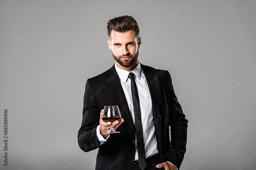 handsome businessman in black suit holding glass with whiskey isolated on grey