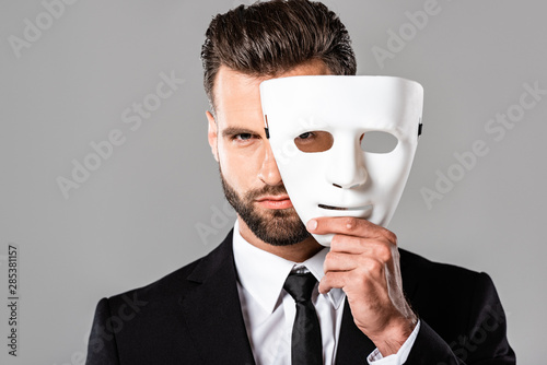 serious handsome businessman in black suit taking off white mask isolated on grey photo