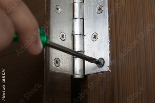 Work's hand uses green screwdriver to install screw on stainless door hinges with wooden swing door for interior design. Horizontal shot.