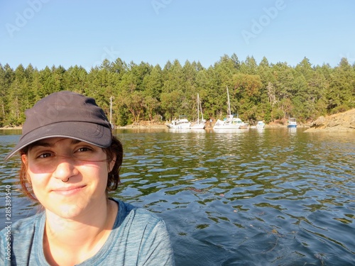A beautiful young woman contently smiling sitting on a small boat surrounded by a beautiful ocean and island full of trees in the background with a few boats behind her on a sunny summer days photo