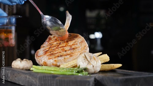 Big juicy grilled steak, Delicious pork, piece of meat served on wooden board with vegetables al dente cooked with flame. Chef decorates dish with spoon putting a pickled tomato. Slow motion foodvideo photo