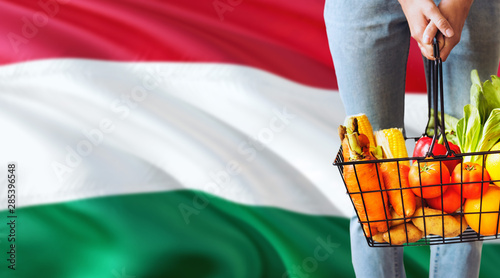 Fototapeta Naklejka Na Ścianę i Meble -  Woman is holding supermarket basket, Hungary waving flag background. Economy concept for fresh fruits and vegetables.