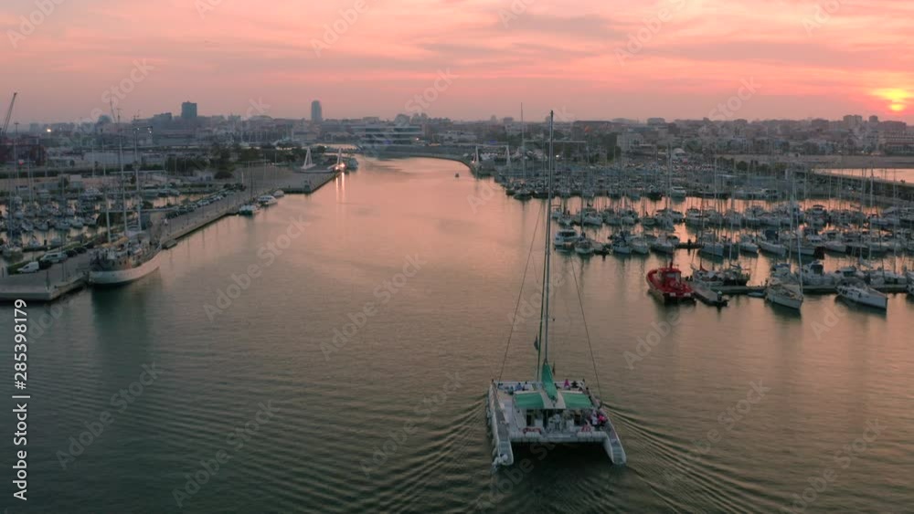A tracking drone shot, following a catamaran sailing through a boat marina, in Valencia, Spain. The sunset's warm colours reflect on the water and the city's horizon line. A tracking drone shot, following a catamaran sailing through a boat marina, in Valencia, Spain. The sunset's warm colours reflect on the water and the city's horizon line.