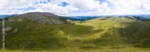 360 degree panoramic aerial view in the wild with green fields and grass and a high mountain with a rocky peak on a warm summer day with white clouds and blue sky with high detail. 3d tour