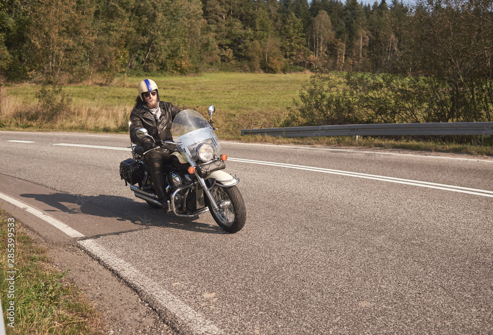 Bearded biker in helmet, sunglasses and black leather clothing riding cruiser bike along sharp turn of empty road on bright summer day on misty background of rural landscape and distant green hills.