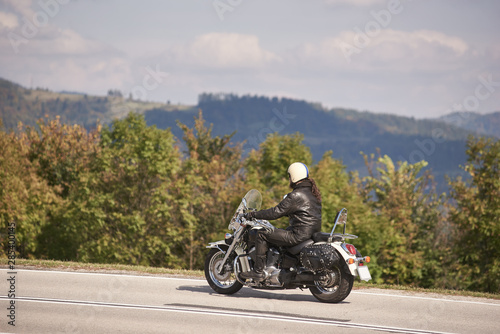 Side view of handsome bearded motorcyclist in helmet  sunglasses and black leather clothing riding cruiser motorbike along narrow asphalt path on sunny summer day.
