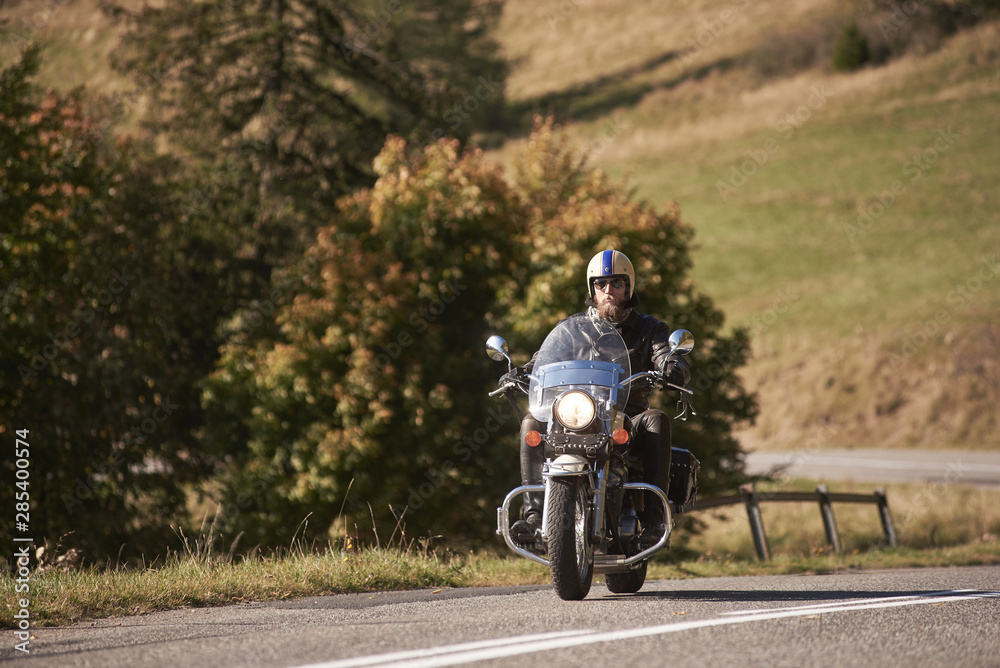 Obraz premium Bearded handsome motorcyclist in helmet, sunglasses and black leather clothing riding cruiser motorbike along empty asphalt road on bright sunny day.