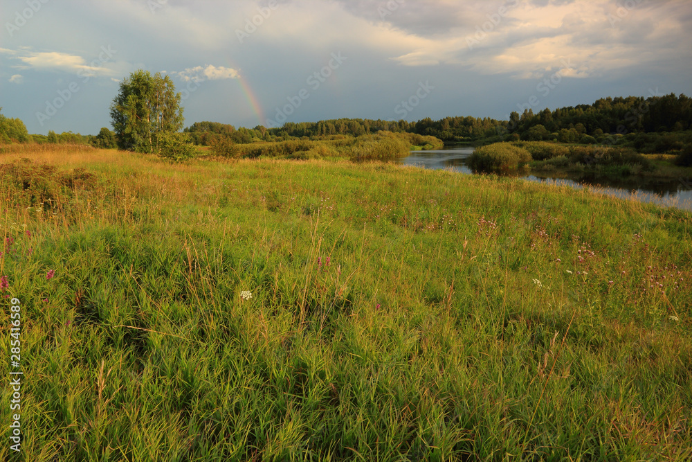Summer evening after rain, rainbow in the sky, view of the riverbed from the high bank.