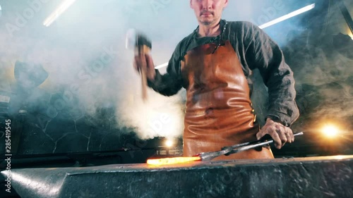 Forger hits hot knife with a hammer on anvil. Blacksmith forging iron in workshop.