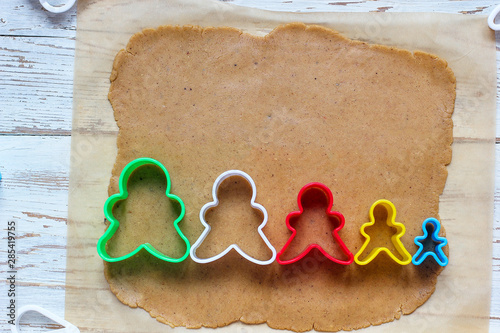 process of dealing with gingerbread man cookies,use red gingerbread man mold cutting gingerbread dough on baking paper around colorful cookie cutters on white wooden table.top view