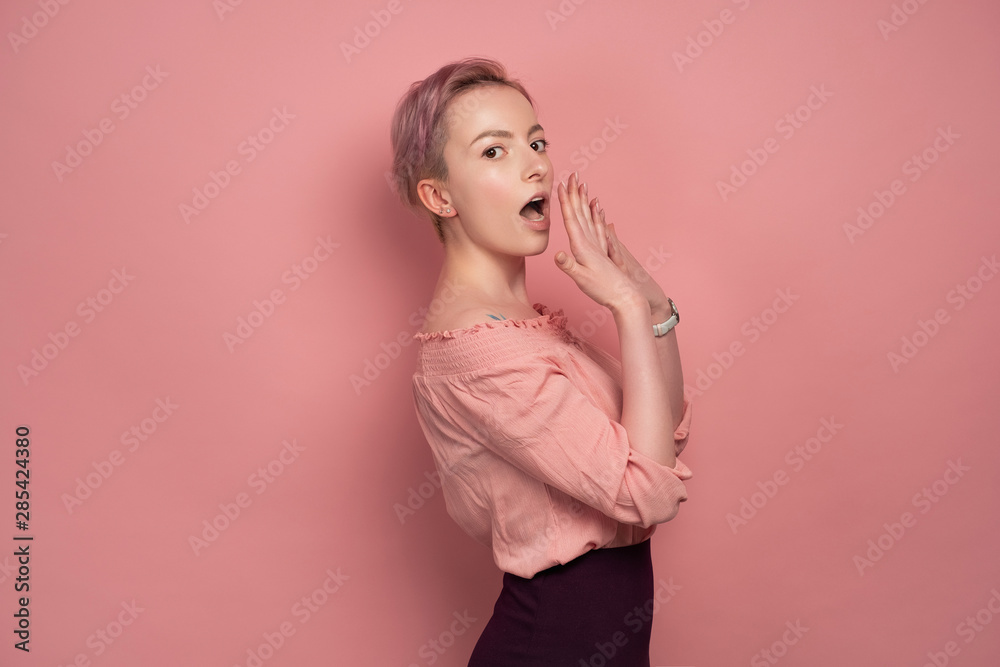 Fototapeta premium A girl with short hair is standing in profile on a pink background and looking at the camera with an embarrassed cover for mouth