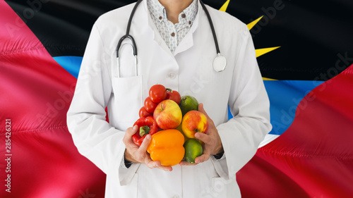 Doctor is holding fruits and vegetables in hands with Antigua and Barbuda flag background. National healthcare concept, medical theme.