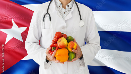 Doctor is holding fruits and vegetables in hands with Cuba flag background. National healthcare concept, medical theme.
