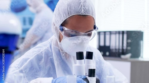 Close up footage of female scientist looking through moden microscope in research laboratory. Scientist in coverall protection equipment. photo