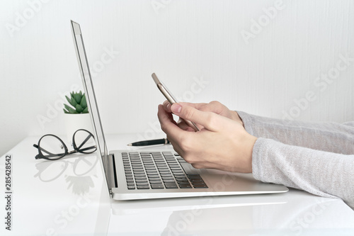 Unrecognizable woman using smartphone while typing on laptop, concept of password entry