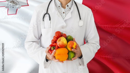 Doctor is holding fruits and vegetables in hands with Malta flag background. National healthcare concept, medical theme.