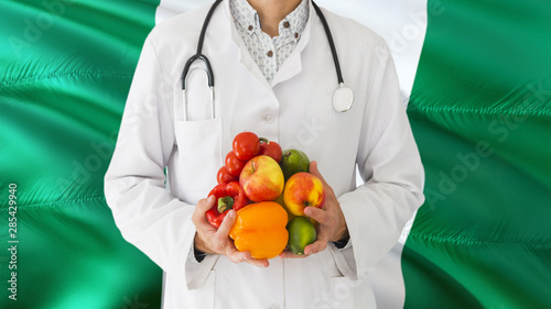 Doctor is holding fruits and vegetables in hands with Nigeria flag background. National healthcare concept, medical theme.