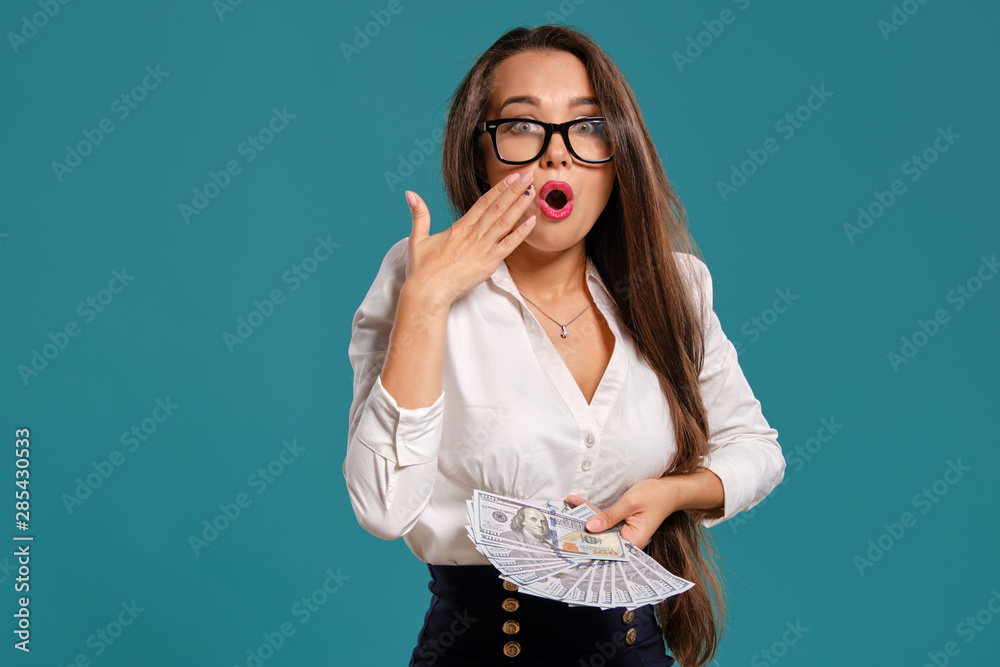 Brunette girl in glasses, wearing in a black short skirt and white blouse is posing holding a fan of hundred dollar bills against a blue background. Brunette girl in glasses, wearing in a black short skirt and white blouse is posing holding a fan of hundred dollar bills against a blue background.