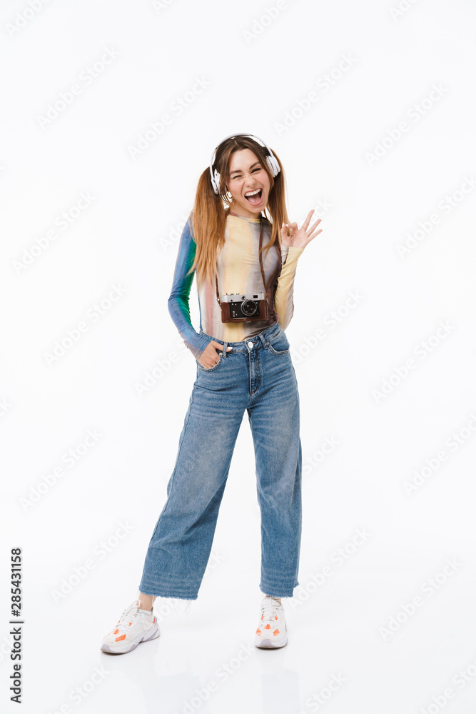Full length photo of cheerful young girl wearing headphones showing ok sign while carrying retro camera Full length photo of cheerful young girl wearing headphones showing ok sign while carrying retro camera