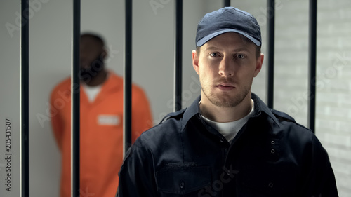 Obraz na plátně Warden looking at camera in prison, african american criminal on background