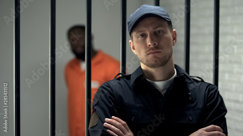 Canvas Print Prison warden looking to camera standing near cell with imprisoned afro-american