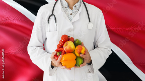 Doctor is holding fruits and vegetables in hands with Trinidad And Tobago flag background. National healthcare concept, medical theme.