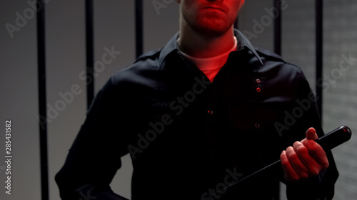 Fotografie Aggressive warden holding truncheon standing against prison cell, red lights