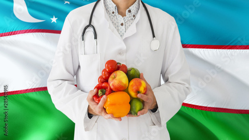 Doctor is holding fruits and vegetables in hands with Uzbekistan flag background. National healthcare concept, medical theme.