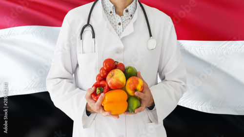 Doctor is holding fruits and vegetables in hands with Yemen flag background. National healthcare concept, medical theme.