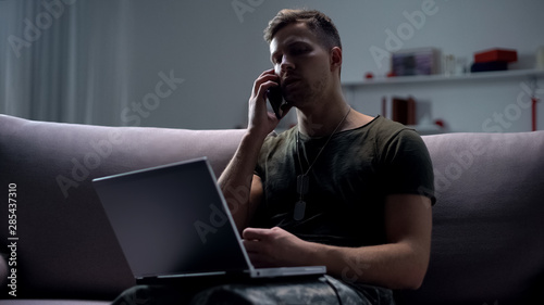Obraz na plátně Male military spy sitting with laptop on sofa and talking smartphone at night