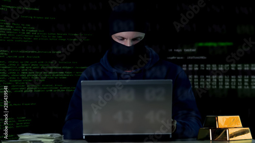 Criminal in balaclava working on laptop, hacking bank security system, data code