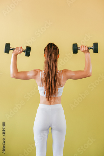 Athletic bodybuilder woman with dumbbells. Beautiful brunette girl with muscles lifting weights on yellow background.