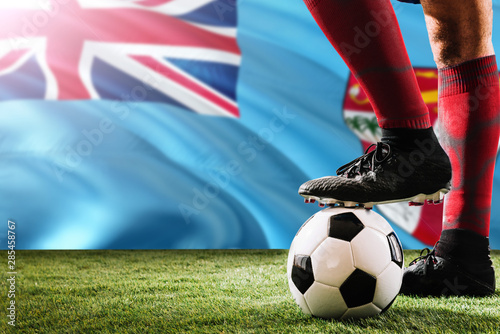 Close up legs of Fiji football team player in red socks, shoes on soccer ball at the free kick or penalty spot playing on grass.