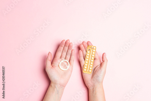 Contraceptive means  a condom and birth control pills in a woman hands on a pink background.