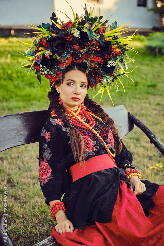 Girl in a white ukrainian authentic national costume and a wreath of flowers is posing sitting on a wooden bench against a white hut. Close-up. Girl in a white ukrainian authentic national costume and a wreath of flowers is posing sitting on a wooden bench against a white hut. Close-up.