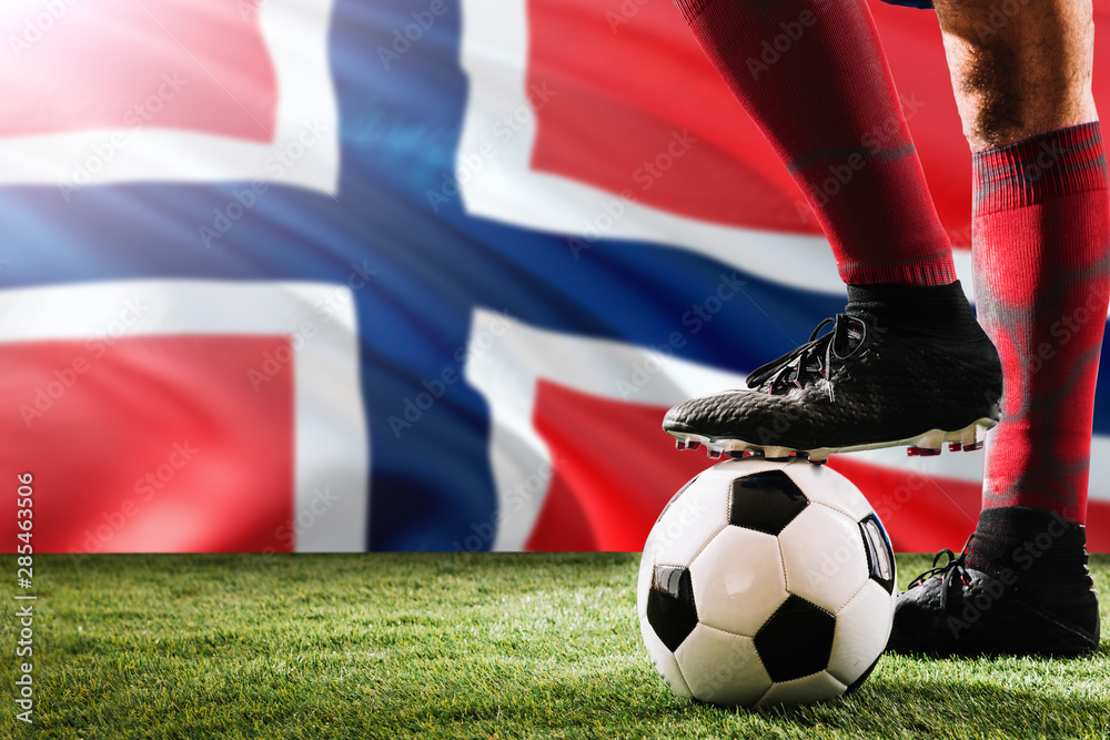 Close up legs of Norway football team player in red socks, shoes on soccer ball at the free kick or penalty spot playing on grass.