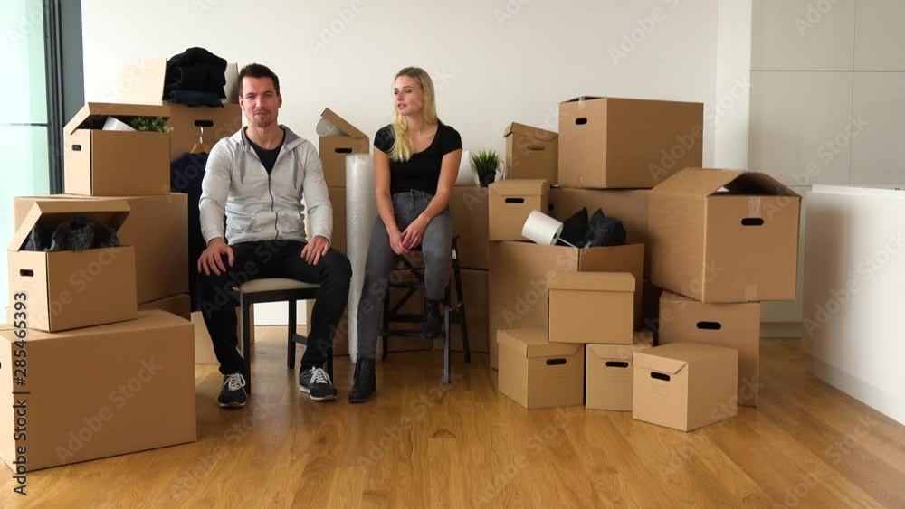 A smiling moving couple sits on chairs and talks to the camera in an empty apartment, surrounded by cardboard boxes A smiling moving couple sits on chairs and talks to the camera in an empty apartment, surrounded by cardboard boxes