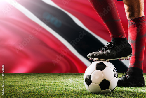 Close up legs of Trinidad And Tobago football team player in red socks, shoes on soccer ball at the free kick or penalty spot playing on grass.