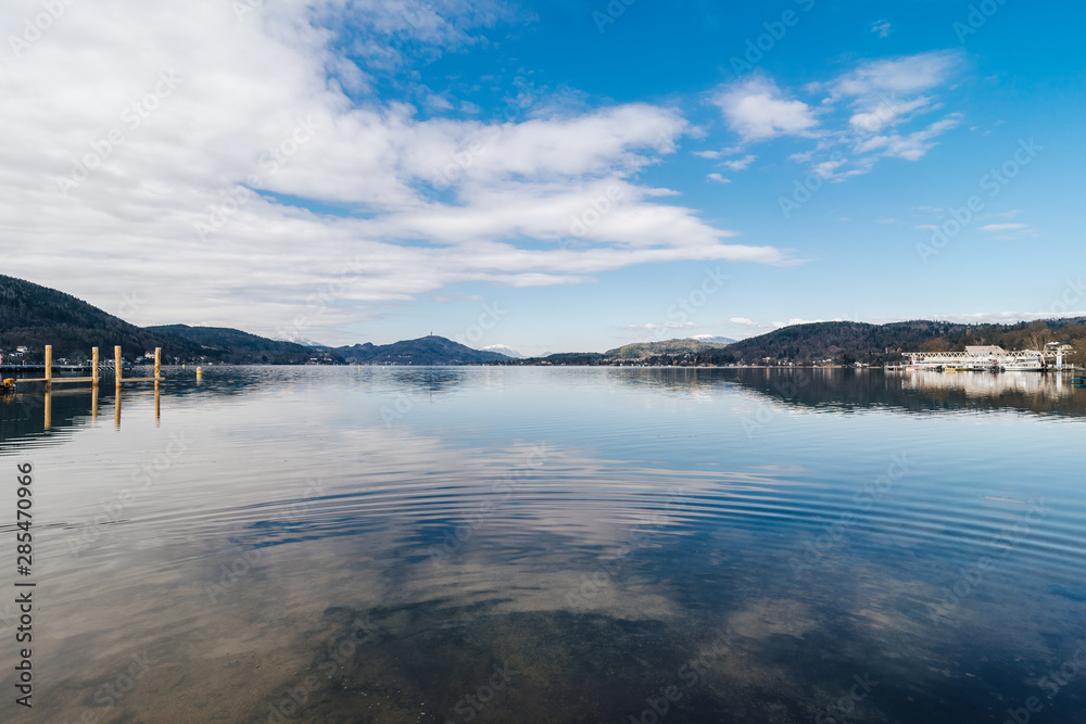 Beautiful view of a famous alpine lake Worthersee, Klagenfurt, Austria.  View of the Worthersee with a pier, clear water and blue sky with white  clouds. Banks of Worthersee lake near Klagenfurt. Stock