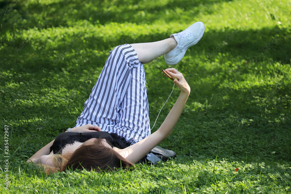 Girl lying on a green grass with smartphone and making selfie. Young woman relaxing in summer park, concept of leisure on nature, watching video, online chat Girl lying on a green grass with smartphone and making selfie. Young woman relaxing in summer park, concept of leisure on nature, watching video, online chat