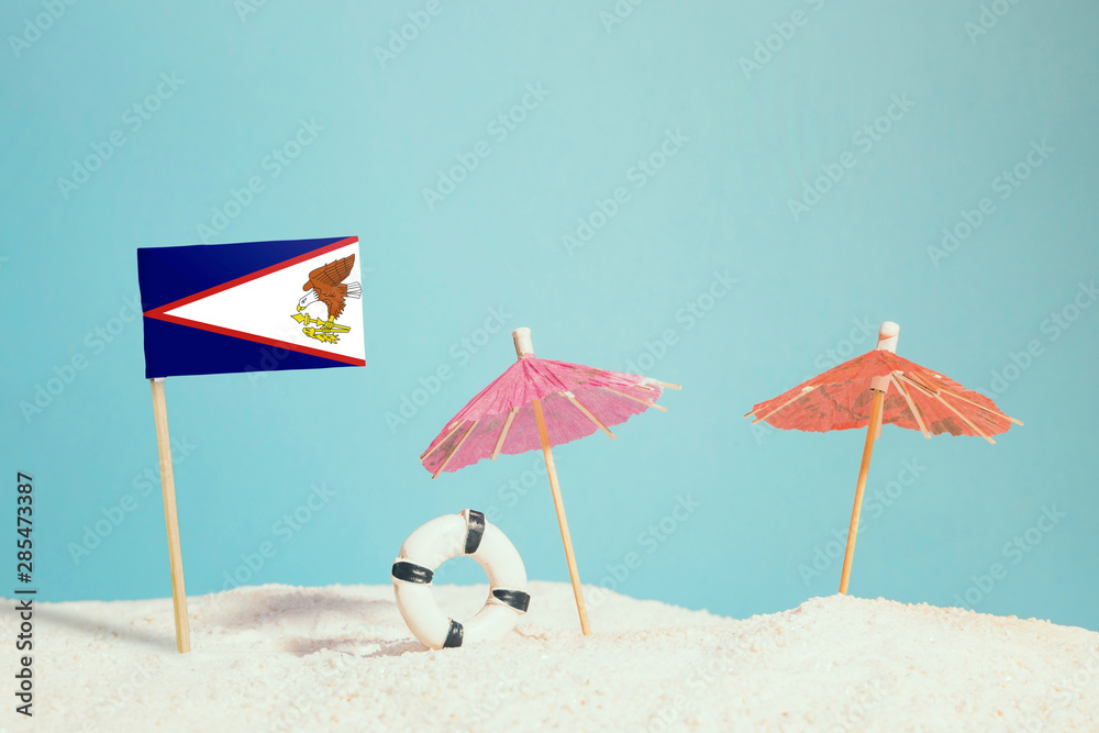 Miniature flag of American Samoa on beach with colorful umbrellas and life preserver. Travel concept, summer theme.
