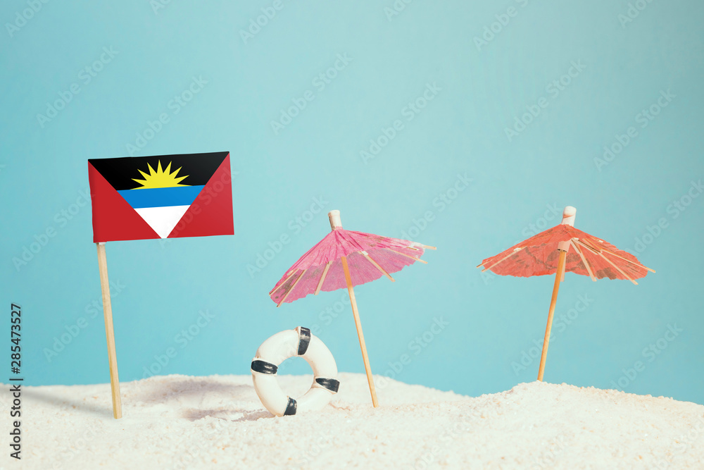 Miniature flag of Antigua and Barbuda on beach with colorful umbrellas and life preserver. Travel concept, summer theme. Miniature flag of Antigua and Barbuda on beach with colorful umbrellas and life preserver. Travel concept, summer theme.