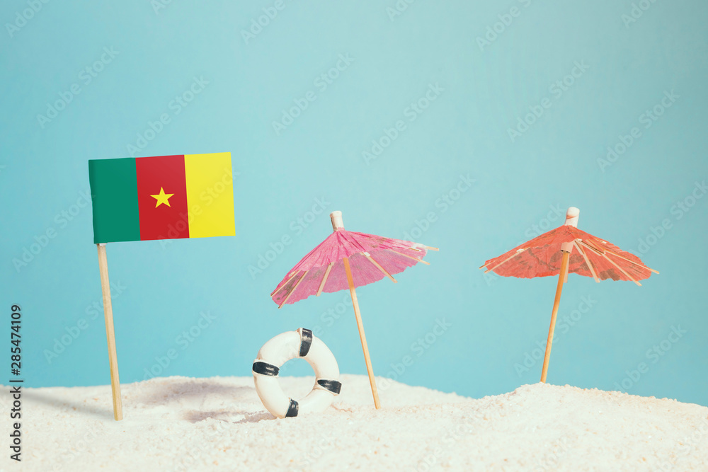 Fototapeta premium Miniature flag of Cameroon on beach with colorful umbrellas and life preserver. Travel concept, summer theme.