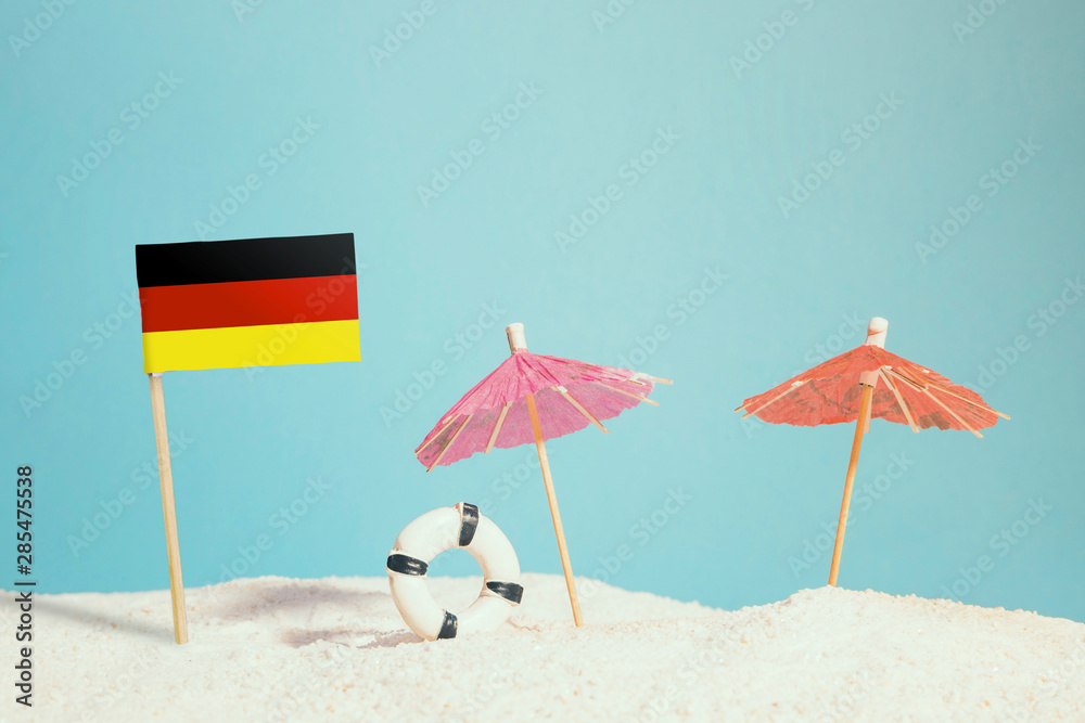 Miniature flag of Germany on beach with colorful umbrellas and life preserver. Travel concept, summer theme.