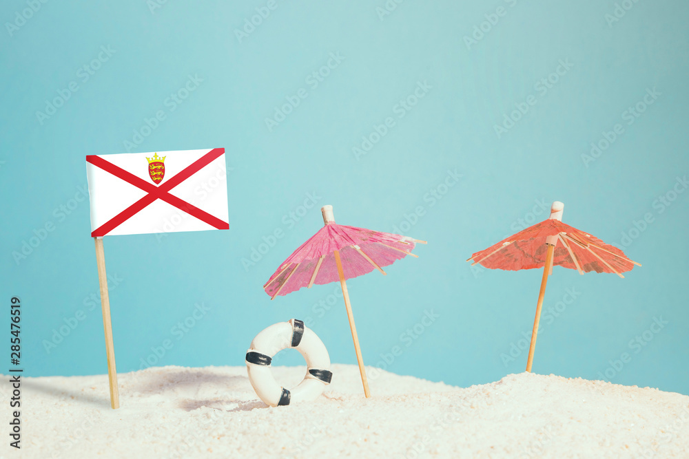 Miniature flag of Jersey on beach with colorful umbrellas and life preserver. Travel concept, summer theme.