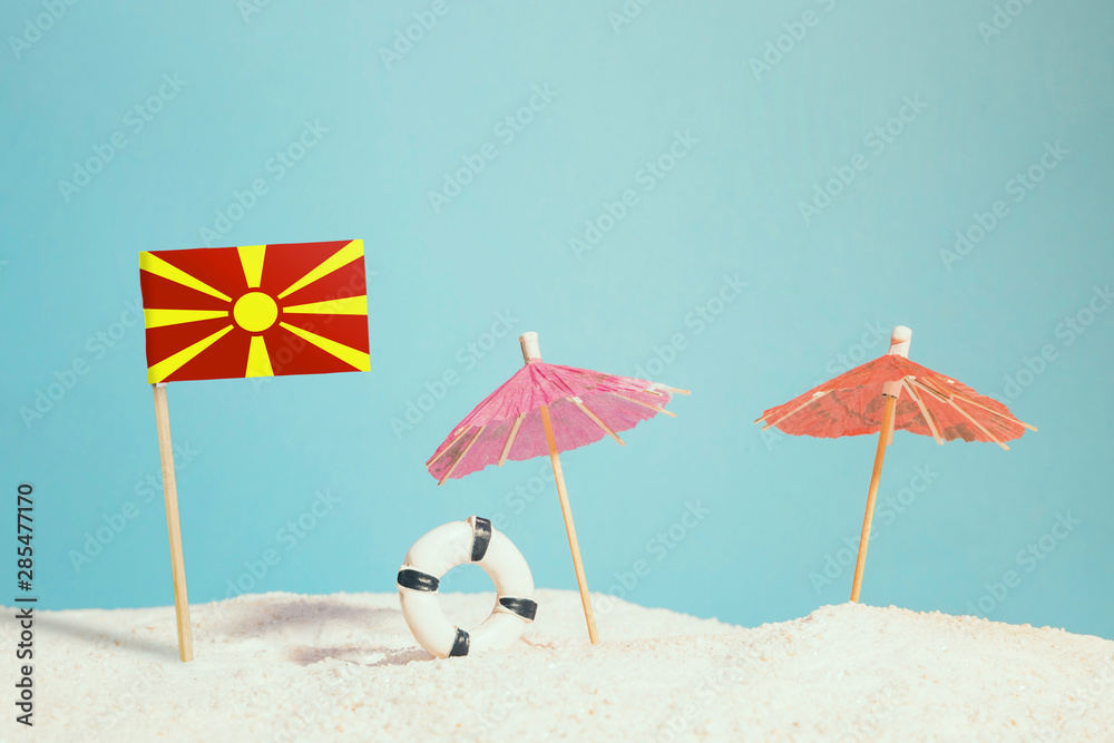 Miniature flag of Macedonia on beach with colorful umbrellas and life preserver. Travel concept, summer theme.