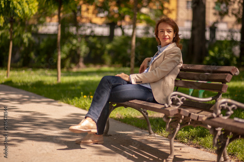 one mature woman, sitting in a bench in a public park outdoors. looking sideways.